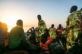 Geflüchtete Menschen die mit Schwimmfesten und Rettungsdecken auf einem Rettungsboot sitzen und stehen. Im Hintergrund der Sonnenuntergang.