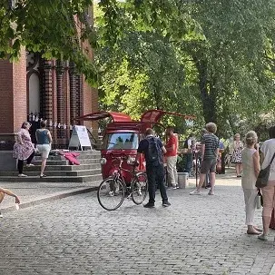 Eine rote Gastro-Ape steht am Rand einer Straße umgeben von grünen Bäumen und mehreren Menschen.