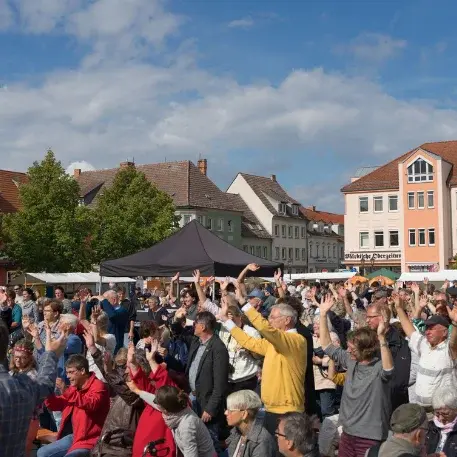 Menschenmenge, die auf einem öffentlichen Platz versammelt ist und in eine Richtung schaut. Viele von ihnen strecken ihre Hände in die Luft.