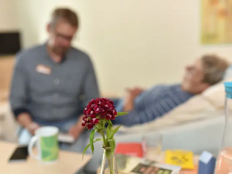 Im Vordergrund steht eine rote Blume in einem Glas. Im unscharfen Hintergrund sitzt ein Mann am Krankenbett einer Frau und liest ihr etwas vor.