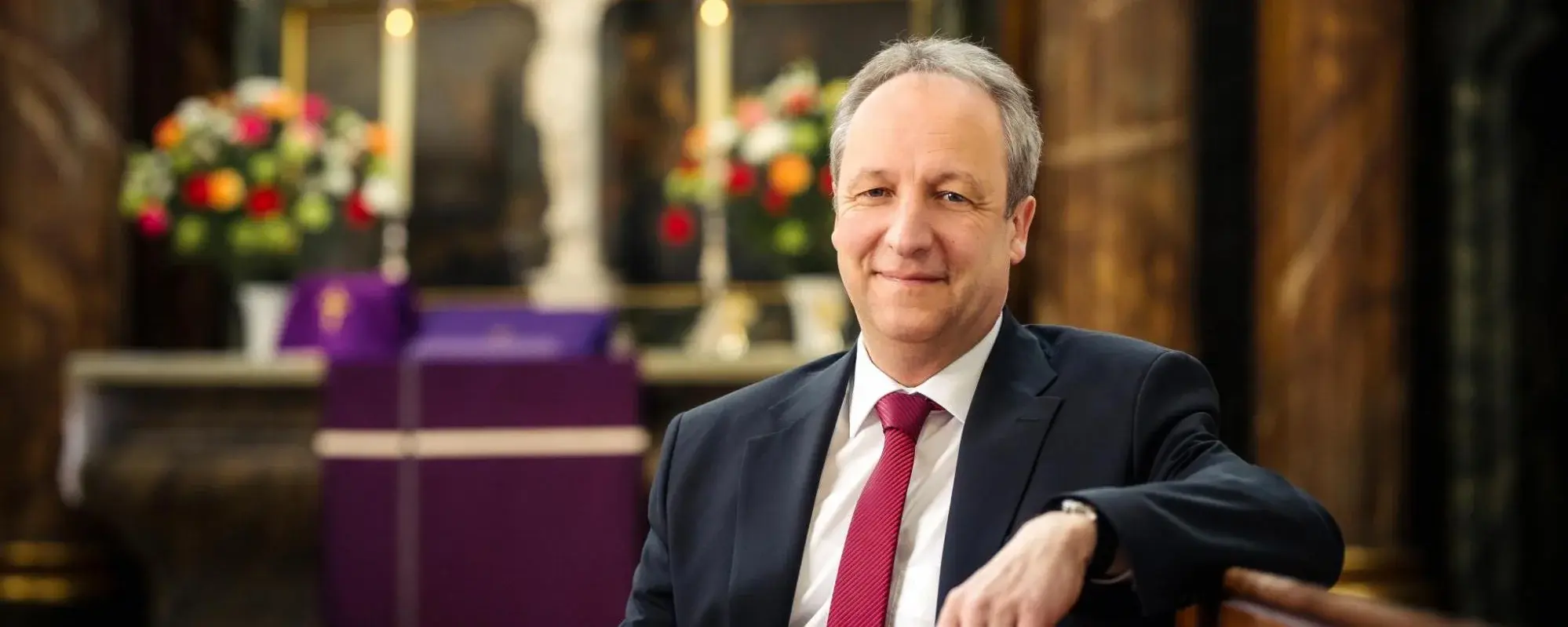 Bischof Stäblein sitzt in einer Kirchenbank und hat den Arm darauf abgelegt. Er trägt einen schwarzen Anzug und lächelt in die Kamera. Im Hintergrund ist verschwommen eine Kirche mit Altar, Blumen und Kruzifix zu sehen.