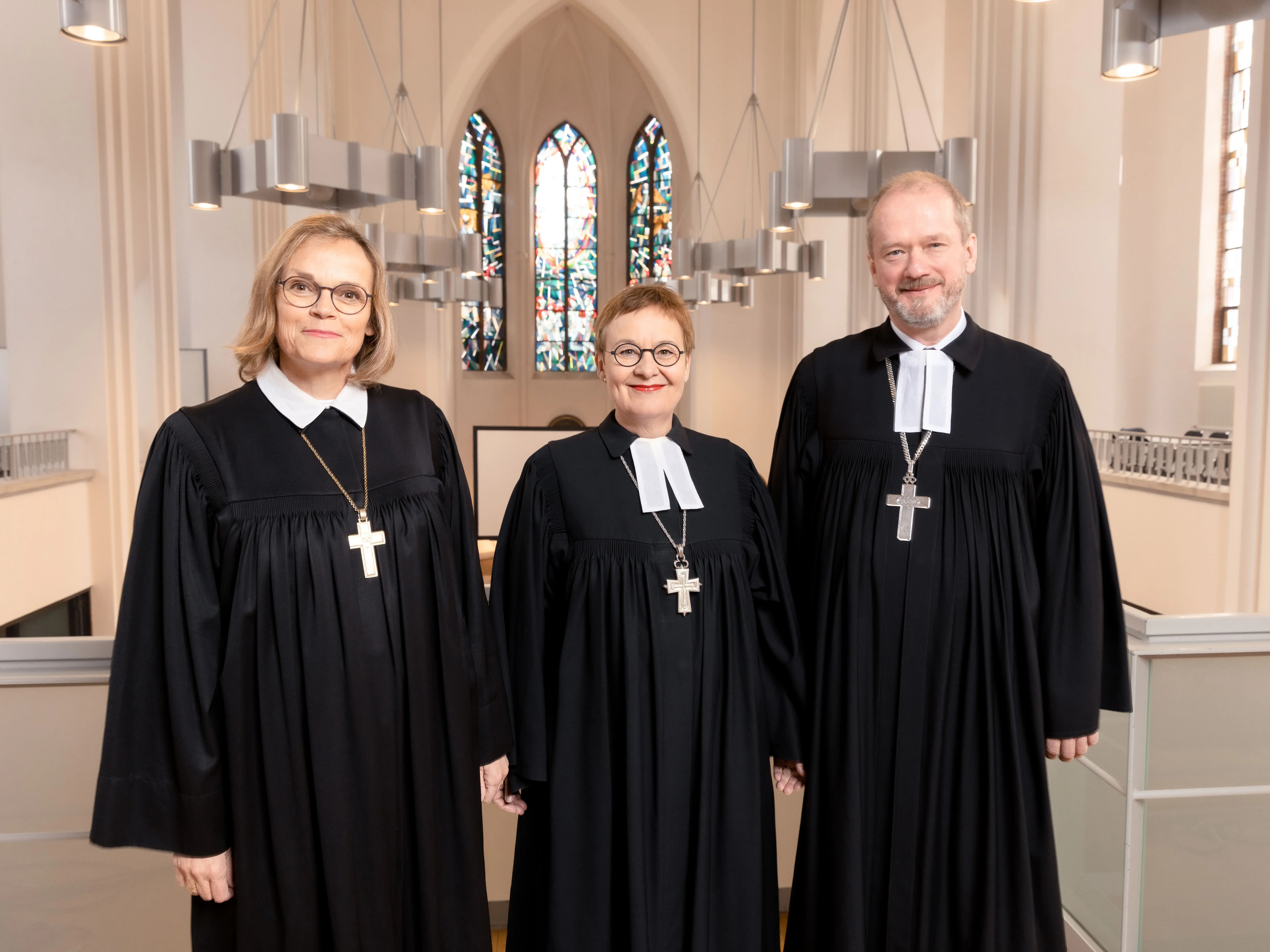 Die drei Generalsuperintendentinnen und Generalsuperintendenten stehen im Talar in einer Kirche und lächeln in die Kamera.