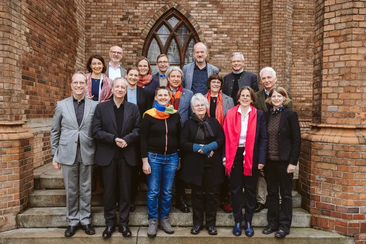 Die Mitglieder der Kirchenleitung auf einer Treppe vor einer Kirchenwand mit Fenster. Alle lächeln in die Kamera.