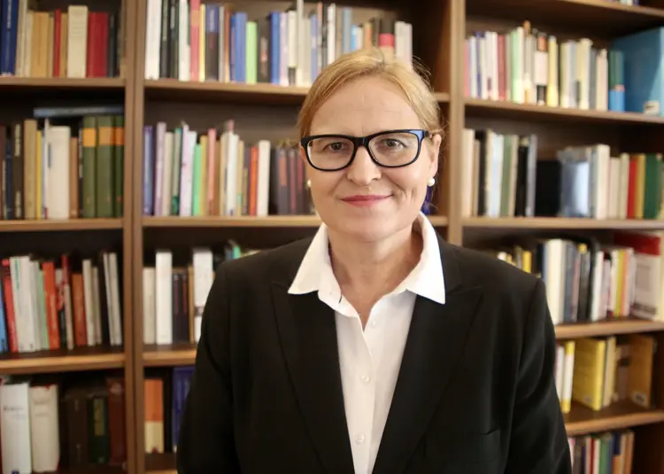 Ulrike E. Auga steht vor einem Bücherregal und schaut in die Kamera.