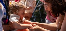 Eine Frau hält die Hände eines kleinen Mädchens, das von einer Person gehalten wird. Sie lächeln sich an.
