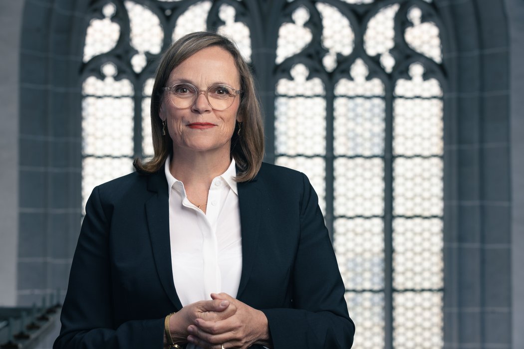 Portrait von Theresa Rinecker in weißer Bluse und schwarzem Blazer. Im Hintergrund ein Kirchenfenster.