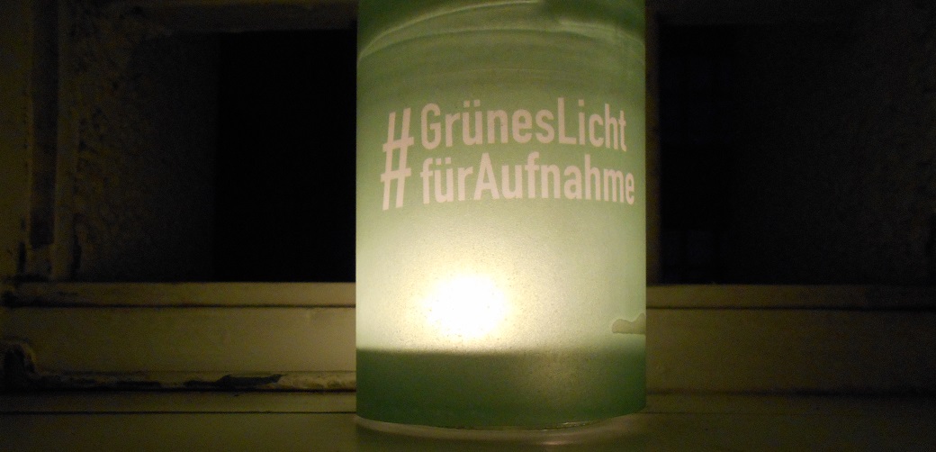 Man sieht ein Kerzenlicht im grünumspannten Glas, Aufschrift: "#Grünes Licht für Aufnahme" vor einem vergitterten Fenster im Dunkeln