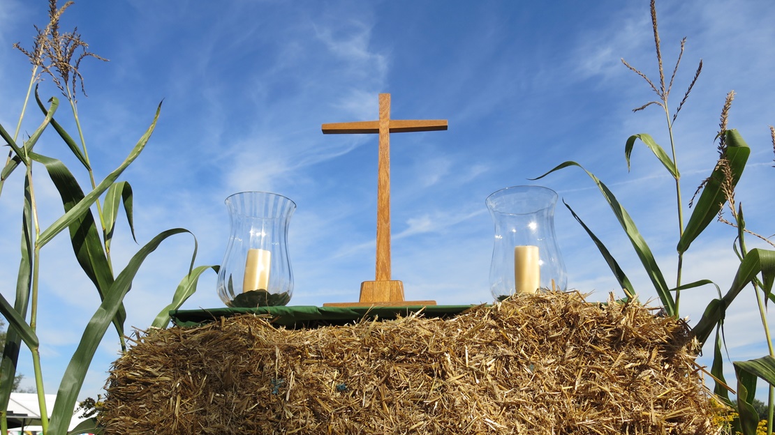 Freiluftaltar: Auf einem Strohballen vor blauem Himmel steht zwischen zwei Kerzen ein hölzernes Kreuz; links und rechts ragt ein Maishalm hervor; Foto: Annette Kaiser/EKBO