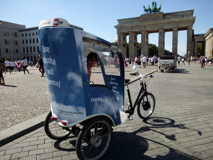 Eine Rikscha voll Himmel, Foto: Alexander Höner. Zu sehen ist eine blaue Rikscha vor dem Brandenburger Tor in Berlin